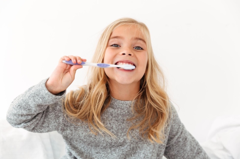 Close-up portrait of cute kid in gray pajamas brushing her teeth, looking at camera
